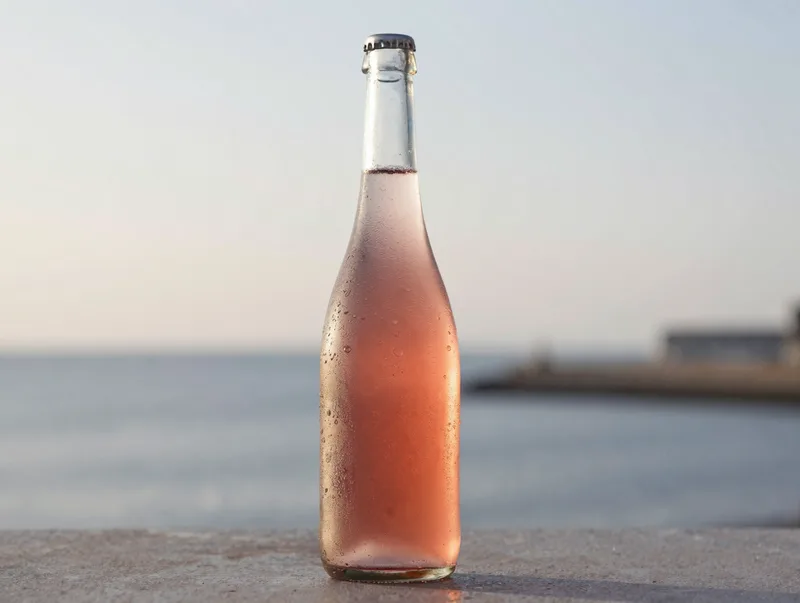 A chilled bottle of pet-nat rose with condensation, pale pink, harbourside evening light