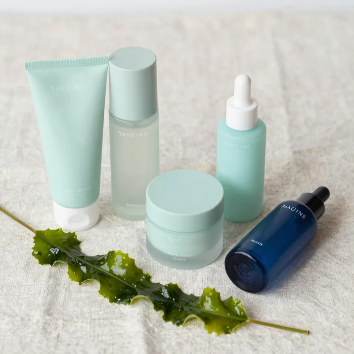 A flat-lay of the full skincare set on pale linen with a sprig of seaweed