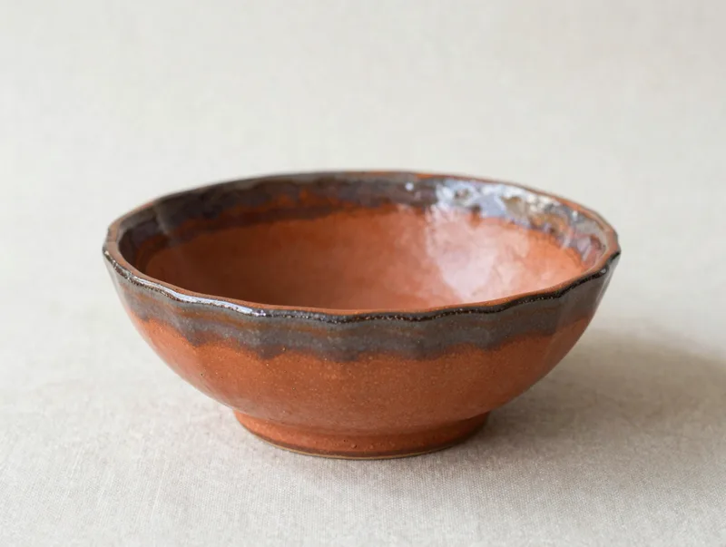 A handmade stoneware ripple bowl in warm terracotta glaze on a linen surface, artisanal ceramics