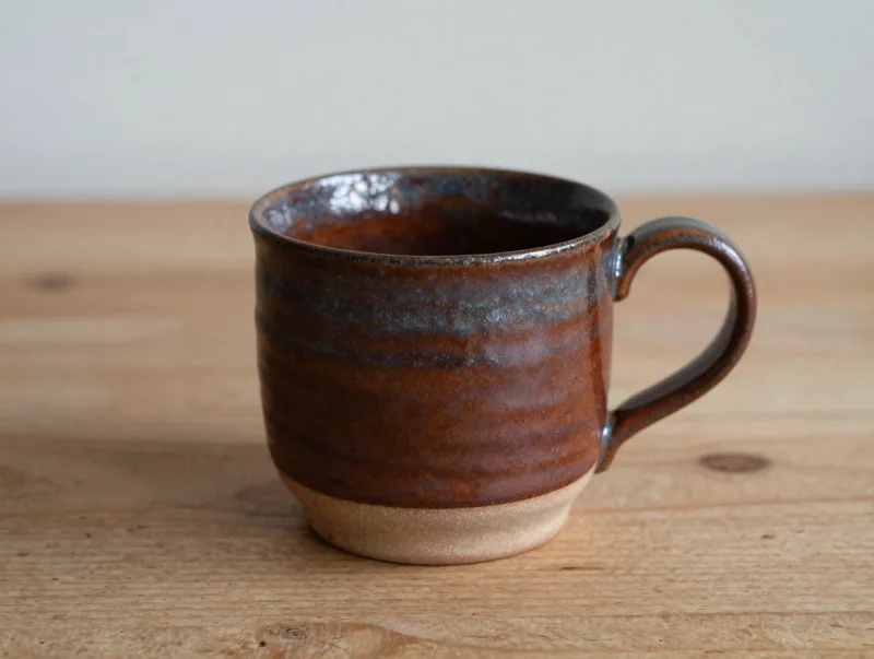A rustic hand-thrown ceramic mug in earthy rust glaze on a wooden table, cozy and tactile