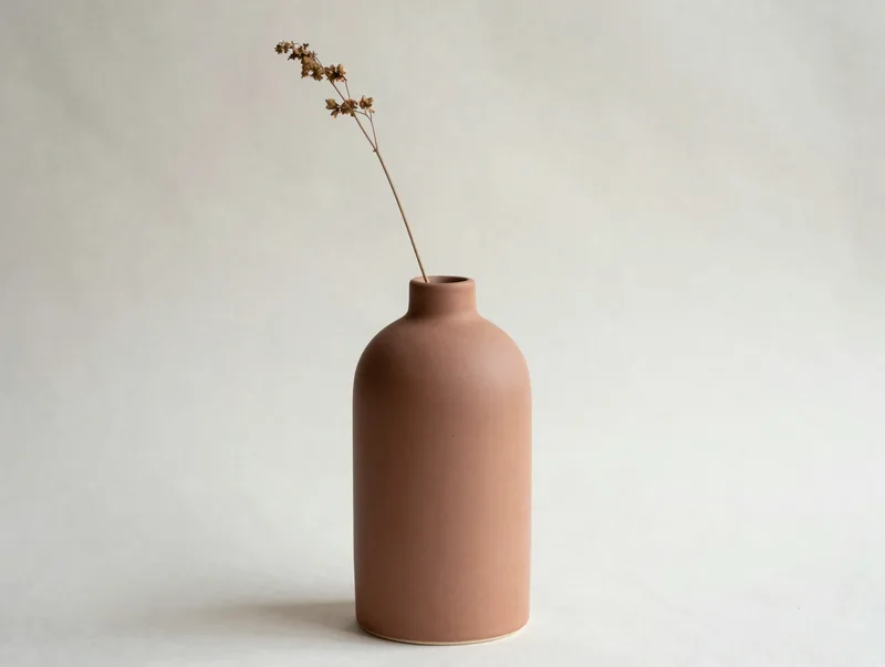 A tall matte ceramic vase in dune tones with a single dried stem, minimal studio still life