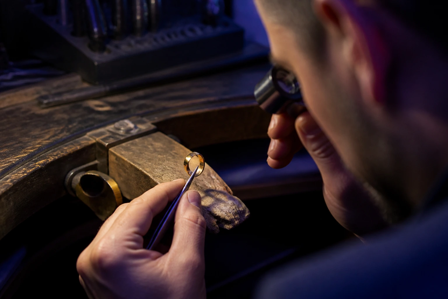 A jeweller's bench in low warm light, loupe, gravers and a single gold ring on a leather pad